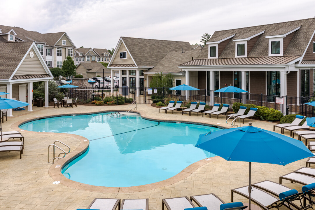 Outdoor pool courtyard with blue umbrellas, lounge chairs, and clubhouse facilities in a resort-inspired setting.