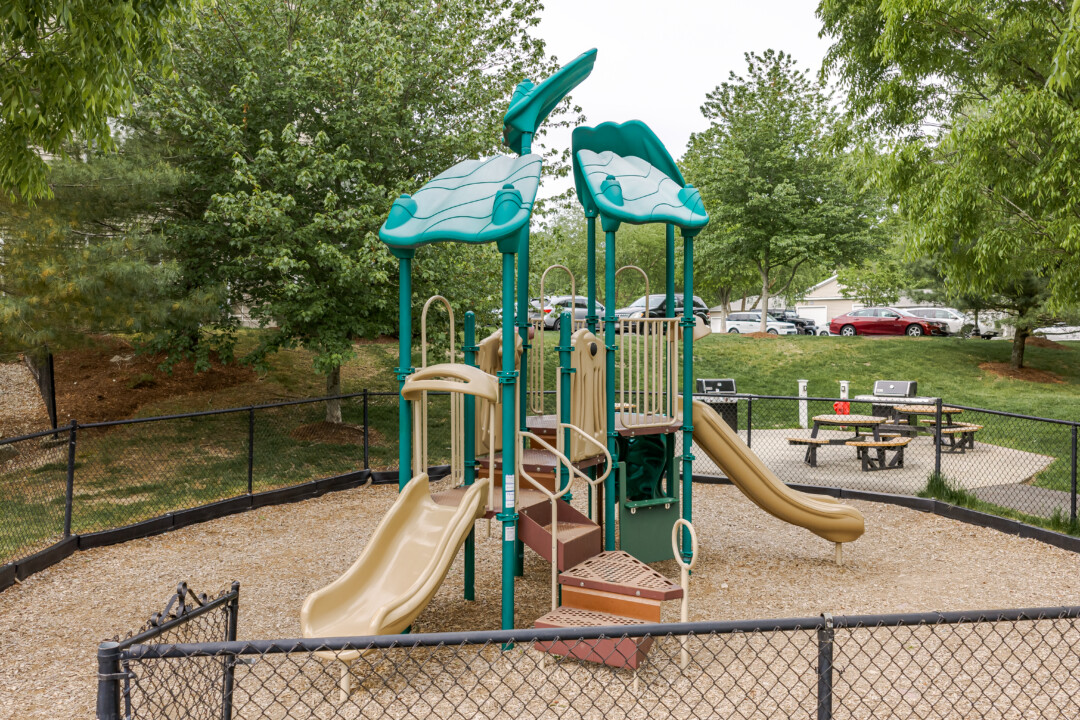 Playground with green and beige play equipment, mulch ground cover, black fencing, and surrounding trees.