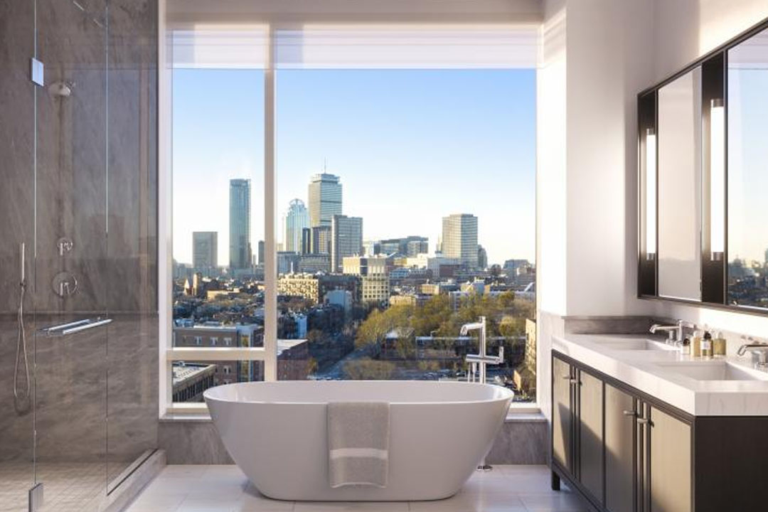 A beautiful tub sits in front of floor-to-ceiling windows and skyline views of the prudential building in Boston. Large walk-in glass shower to the left.