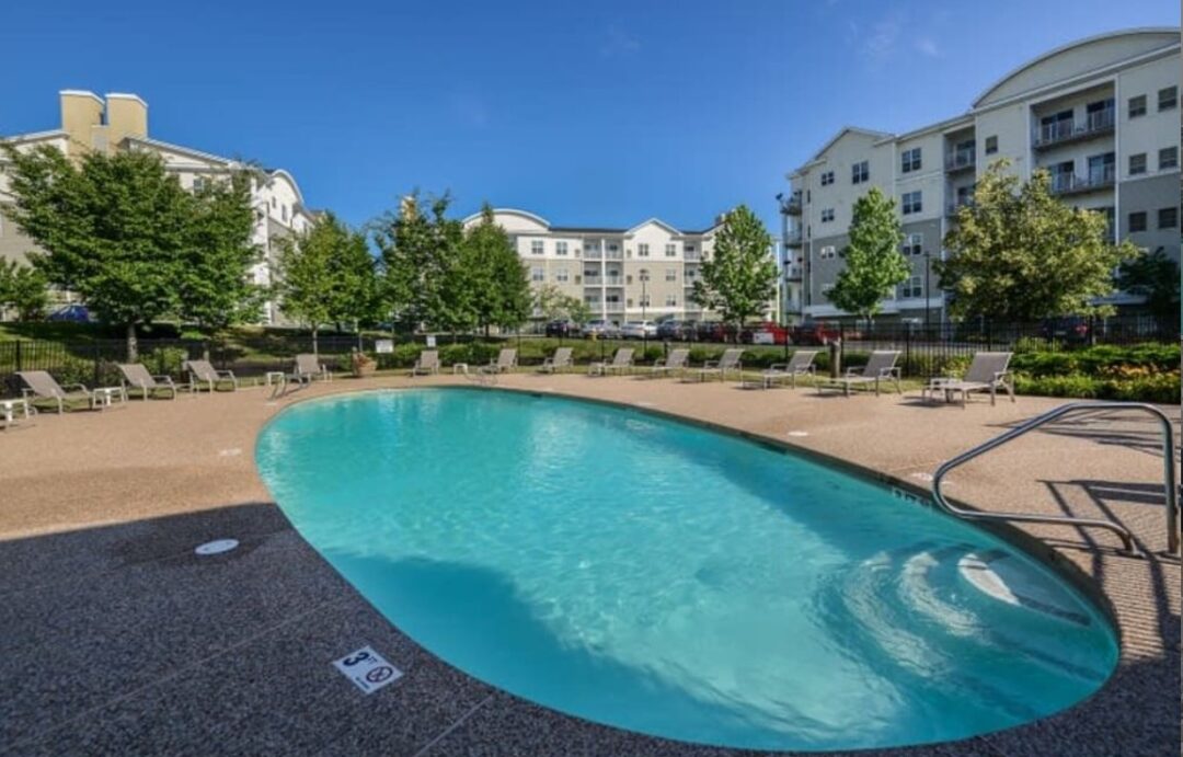 A large, resort-style swimming pool at an apartment complex with lounge chairs, umbrellas, and surrounding landscaping