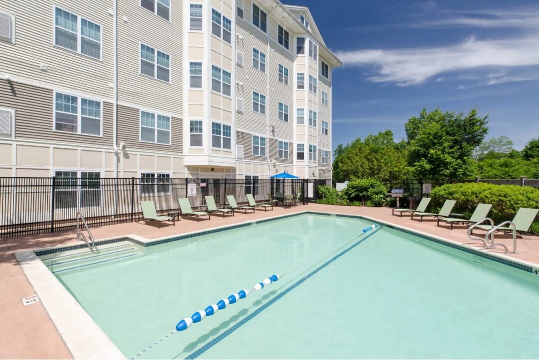 A large, resort-style swimming pool at an apartment complex with lounge chairs, umbrellas, and surrounding landscaping