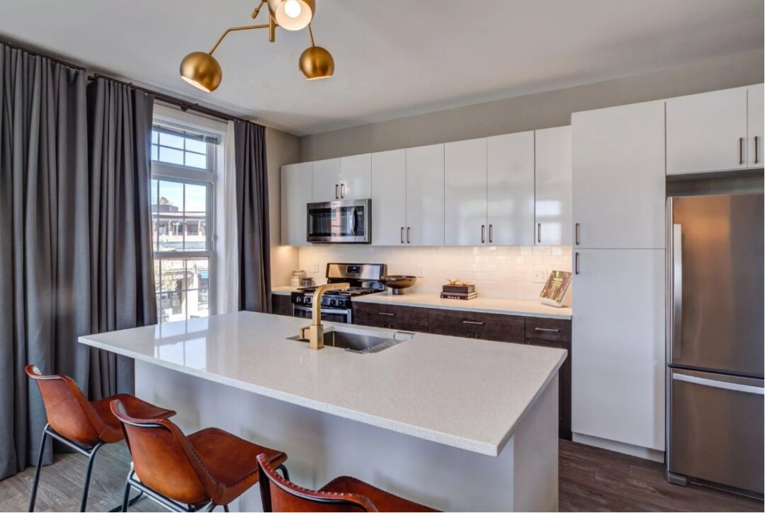 A modern, open-concept kitchen featuring Caesarstone quartz countertops, dark wood cabinetry, and a full suite of stainless steel appliances. The space includes a large center island with pendant lighting, a tile backsplash, and wide-plank wood-style flooring that flows into the living area