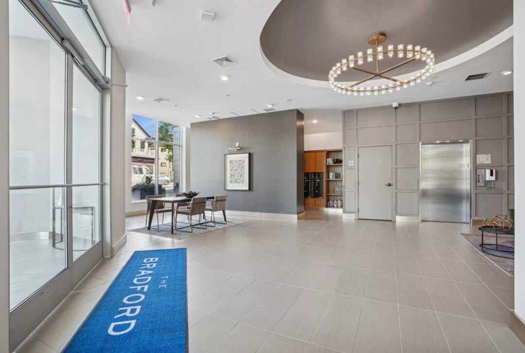A modern, boutique-style apartment lobby featuring floor-to-ceiling windows that provide ample natural light. A mailroom area and shiny silver elevator doors