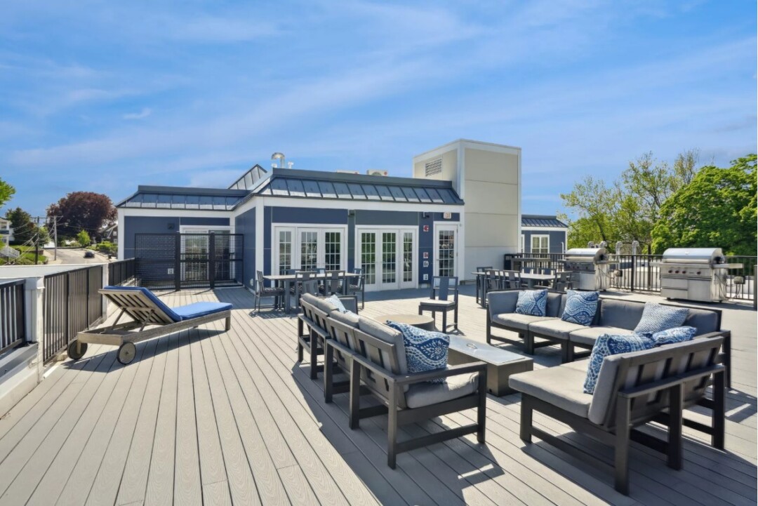 An expansive rooftop sundeck at The Bradford, featuring multiple lounge seating areas, modern outdoor firepits, and stainless steel grill stations. The space is enclosed by a glass railing offering panoramic views