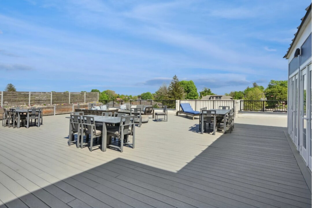 An expansive rooftop sundeck at The Bradford, featuring multiple lounge seating areas, modern outdoor firepits, and stainless steel grill stations. The space is enclosed by a glass railing offering panoramic views