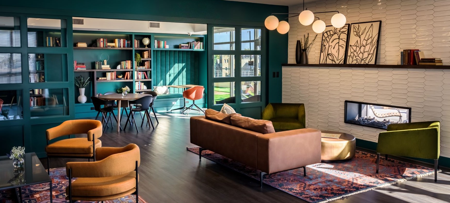 Stylish lounge area with teal built-in shelving filled with books, a seating area with sofa and chairs on a patterned rug, a fireplace set into a tiled wall with a shelf above, and a table near large windows.