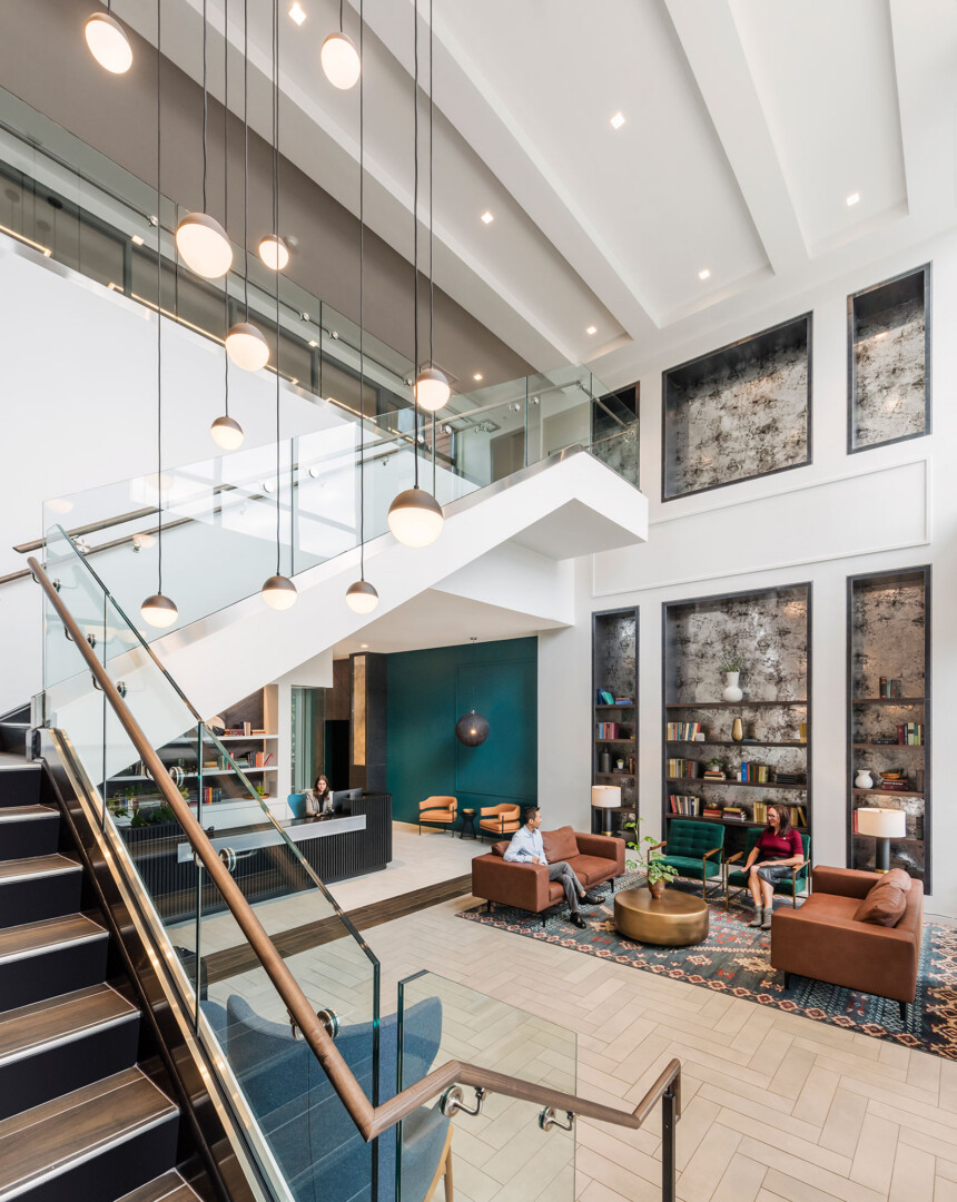 Lobby with high ceilings, a staircase with glass railing, hanging light fixtures, built-in shelving, and a seating area with brown sofas and chairs arranged around a rug.