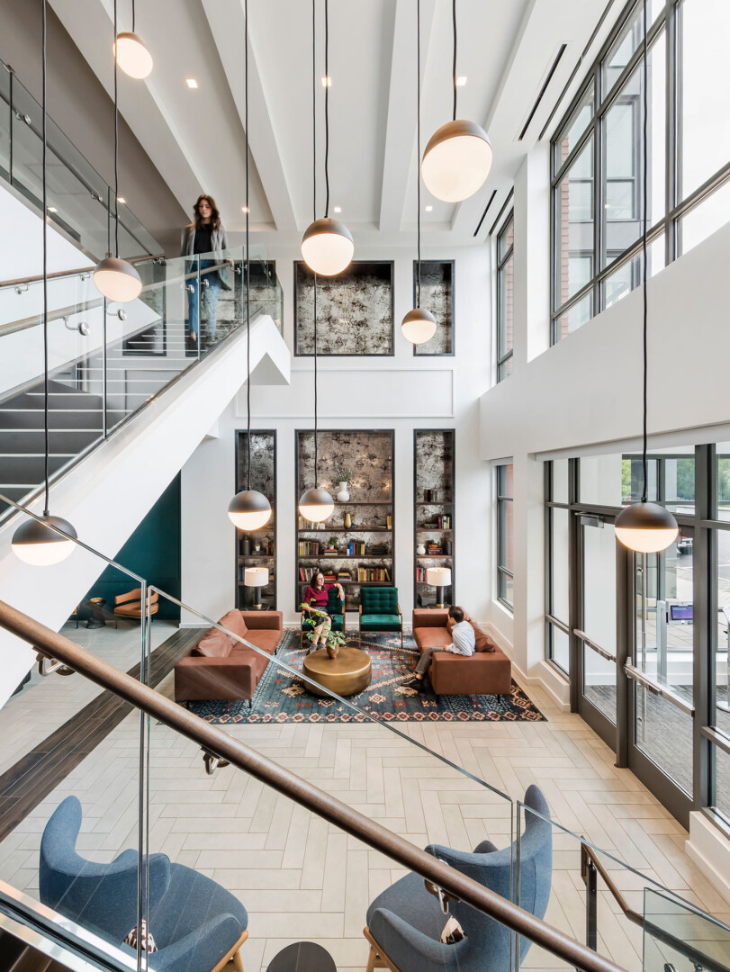 Lobby with high ceilings, a staircase with glass railing, hanging light fixtures, built-in shelving, and a seating area with brown sofas and chairs arranged around a rug.