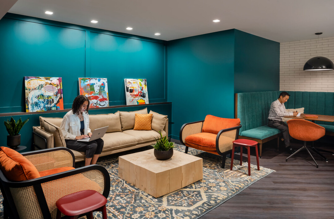 Lounge area with teal walls, a sofa and chairs arranged around a coffee table on a patterned rug, colorful wall art, and a booth-style seating area with a table.
