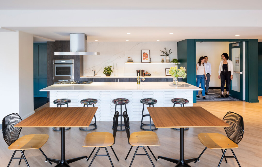 Kitchen and dining area with white walls and ceiling, featuring a large white island with a built-in cooktop and four round bar stools. Behind the island are dark wooden cabinets with a white countertop, a wall oven, a hanging shelf with framed decor and plants, and a white marble wall. Wooden tables with matching chairs are in the foreground. Three people walk through a doorway at the back, leading into the space.