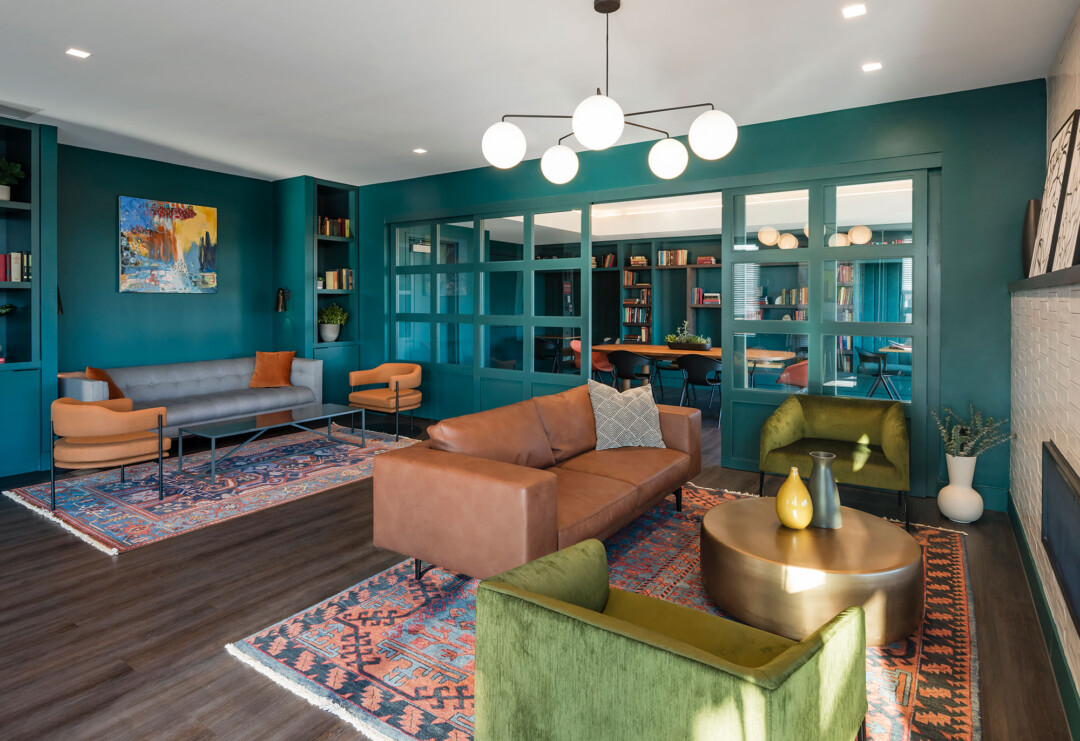 Lounge area with teal walls, sofas and chairs arranged around coffee tables on patterned rugs, built-in shelving, and a glass-enclosed room with a table and seating in the background.