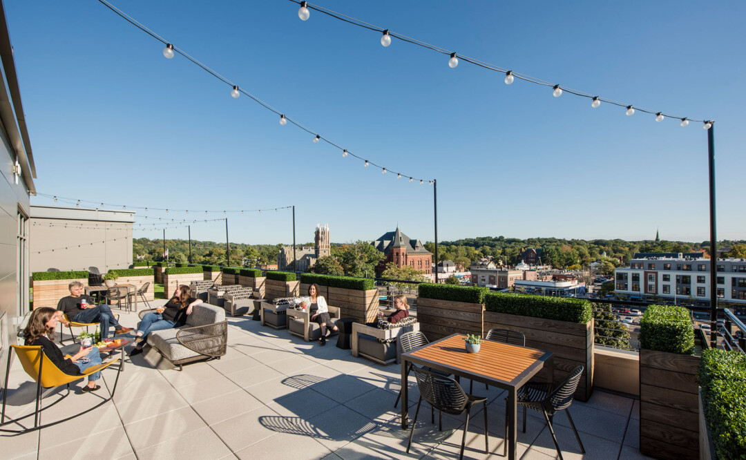 Rooftop terrace with seating areas, tables, and string lights, overlooking a cityscape, with people relaxing and socializing.