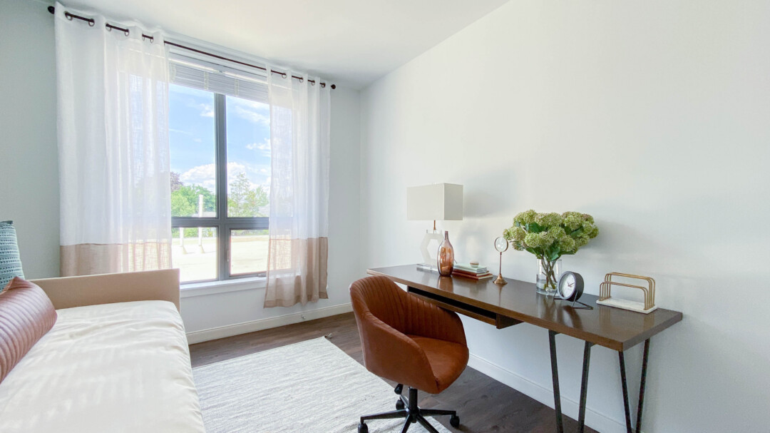 Light-filled bedroom with white walls and ceiling, bed, window with sheer curtains, wooden desk with lamp and décor, and a brown upholstered chair.