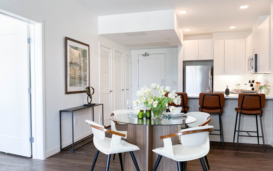 Dining area with white walls, round table and white chairs, white floral centerpiece, wall art, and adjacent kitchen with white cabinets, refrigerator, kitchen bar seating, and brown bar stools.