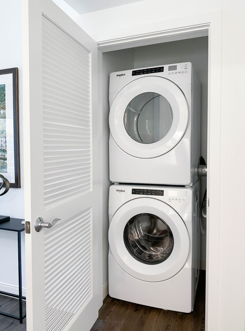 Stacked white front-load washer and dryer in a closet nook with a louvered door, white walls, and dark wood flooring.