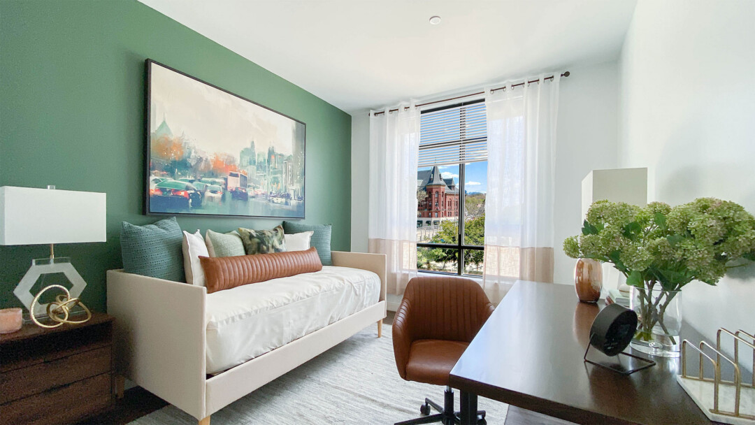 Bedroom with a white bed against a green accent wall, styled with multiple pillows, large framed artwork above, and a wooden nightstand with a lamp. A window with sheer curtains with outside view. A wooden desk with a brown chair sits on the opposite side, decorated with a vase of green flowers and a lamp.