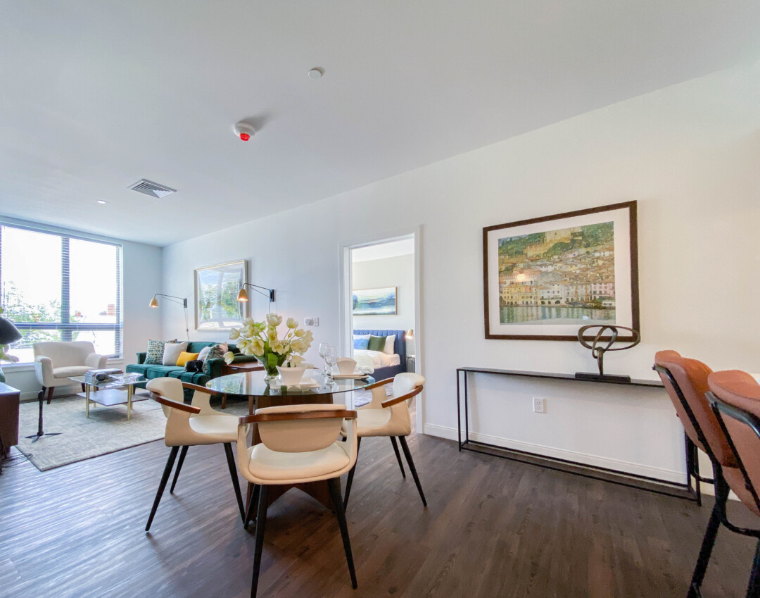 Dining and living area with white walls and ceiling, featuring four upholstered white chairs around a round glass dining table topped with a white floral centerpiece. A console table with a decorative sculpture sits beneath a framed painting on the wall. In the back is a living room with a green sofa, white padded chairs, floor lamps, and a large window that provides natural light, with a bedroom visible through an open doorway.