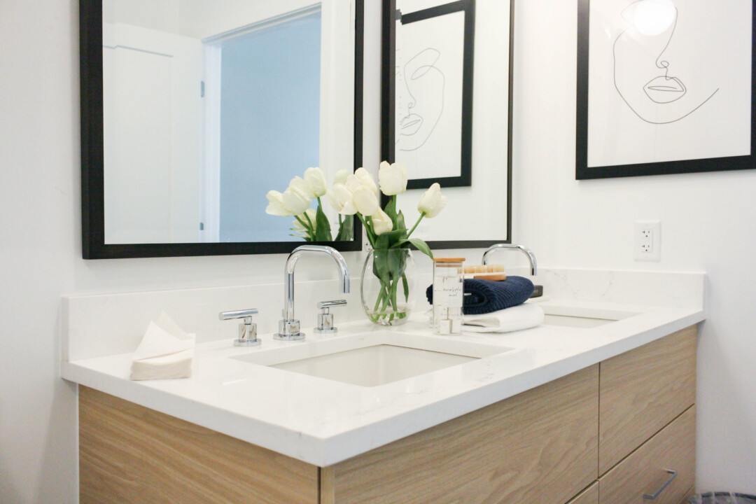 Bathroom with white walls and a double-sink wood vanity with drawers and a white countertop. Two faucets sit beneath large framed mirrors, with artwork on the wall. The countertop is styled with white flowers in a glass vase, folded towels, and small accessories.