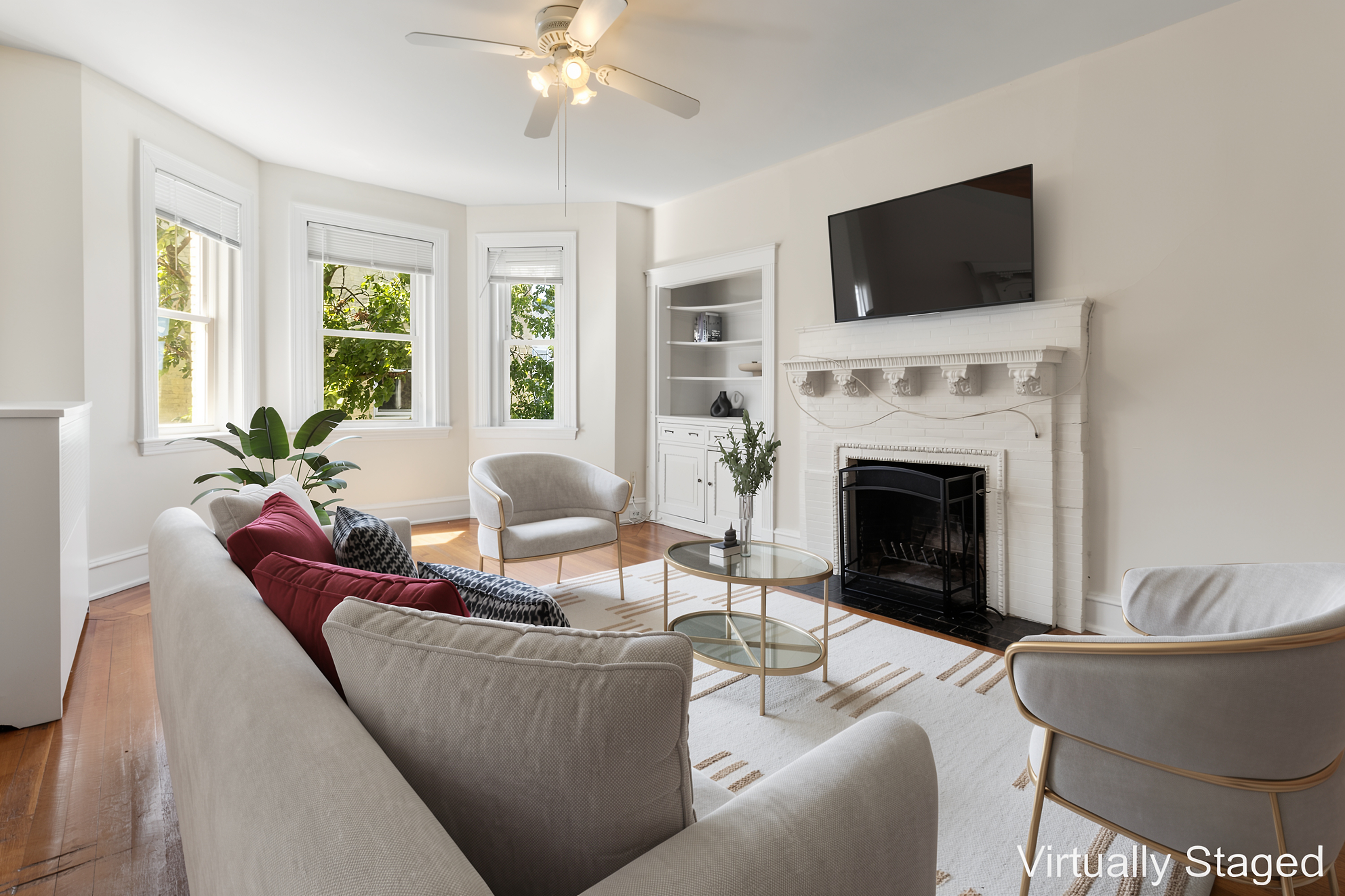 Living room with white walls, hardwood flooring, a decorative fireplace with mantel, multiple windows, a ceiling fan, and with gray sofa, chairs, coffee table, and wall-mounted television.