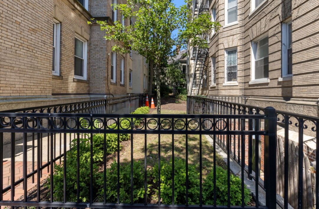 Landscaped outdoor space between two brick apartment buildings with black metal fencing, trees, shrubs, and paved walkway