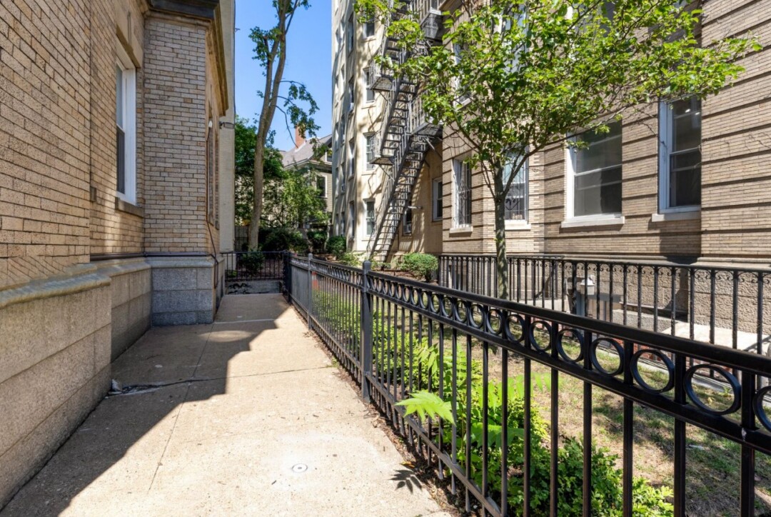 Concrete walkway between two brick apartment buildings with black metal fencing, trees, landscaping, and windows along both buildings.