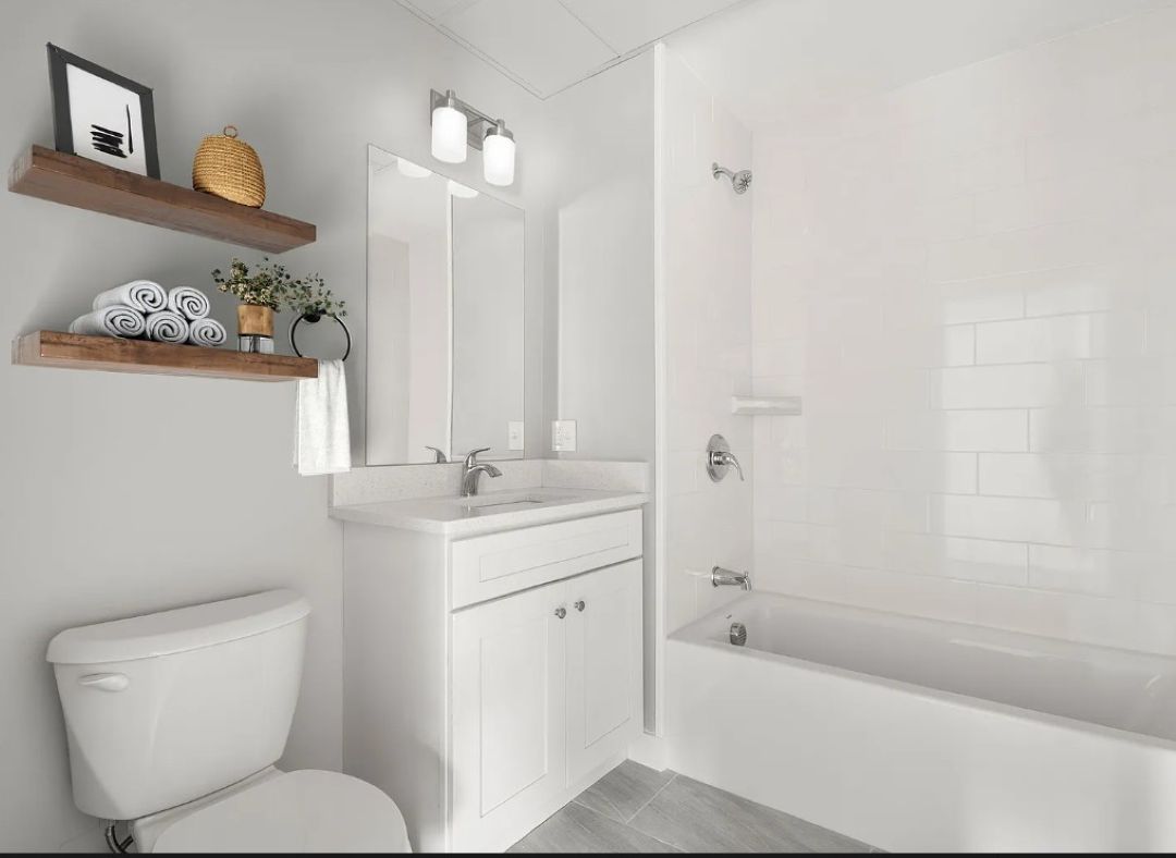 Bathroom with white vanity, white countertop, light gray tile flooring, and a white bathtub with a tiled shower surround. Floating wood shelves and a large mirror.