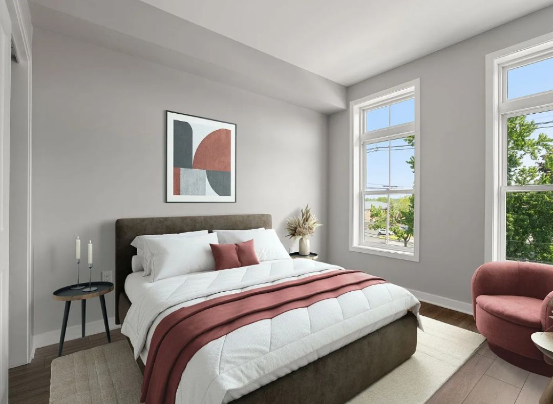 Bedroom with light gray walls, wood-look flooring, and two large windows. Includes a large size bed with white and burgundy bedding, side tables on each side of the bed, a burgundy accent chair, and simple modern décor.