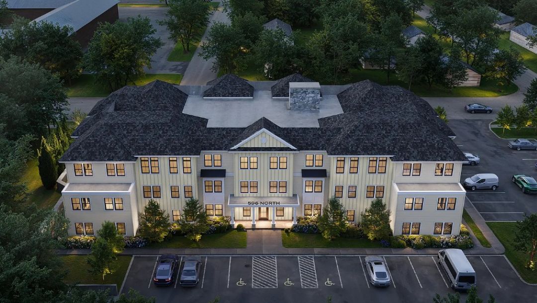 Three-story beige apartment building with white trim and a dark gray roof, illuminated by warm exterior lighting. A paved parking lot with marked spaces sits in front, surrounded by trees.