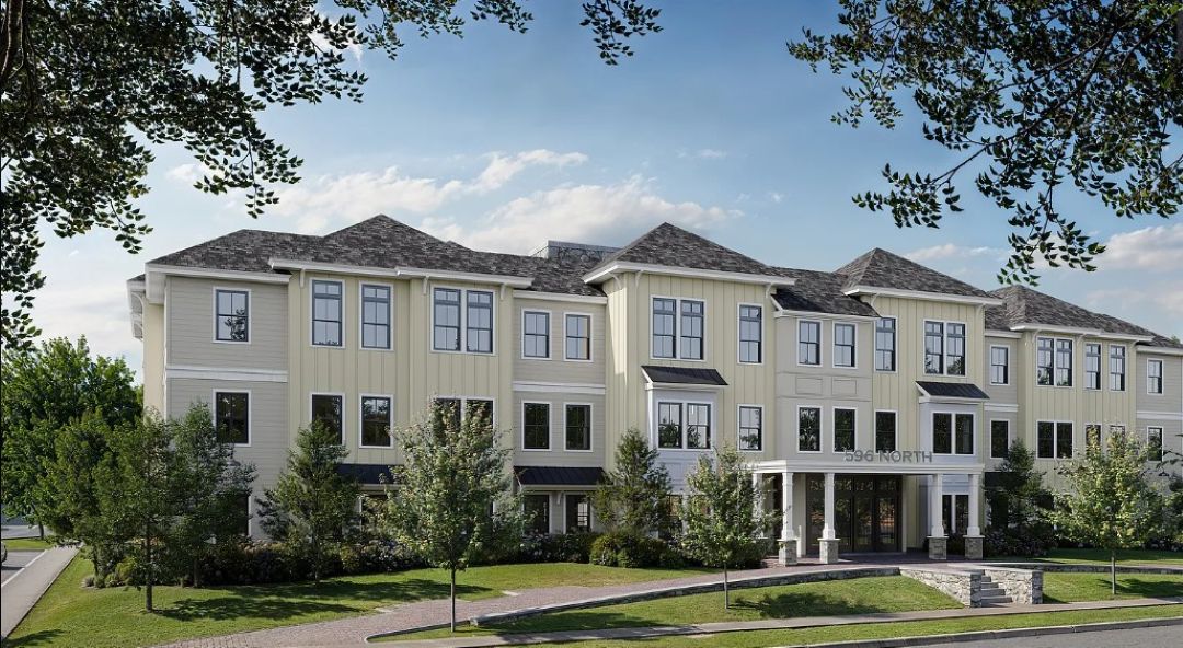 Three-story beige apartment building with white trim, and a dark gray shingled roof. Large rectangular windows and landscaped greenery line the front of the building.