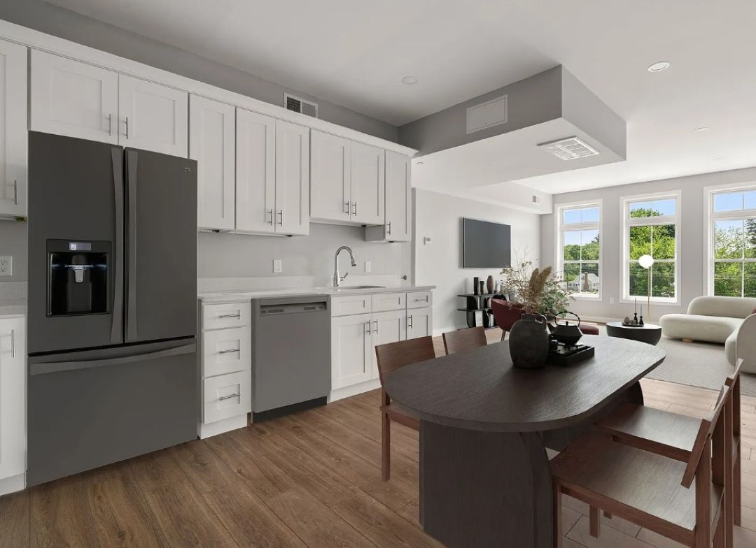 Kitchen with white shaker cabinets, light colored walls, wood-look flooring, and white countertops. A dark wood dining table sits beside a dark colored stainless steel refrigerator, with large windows bringing in natural light.