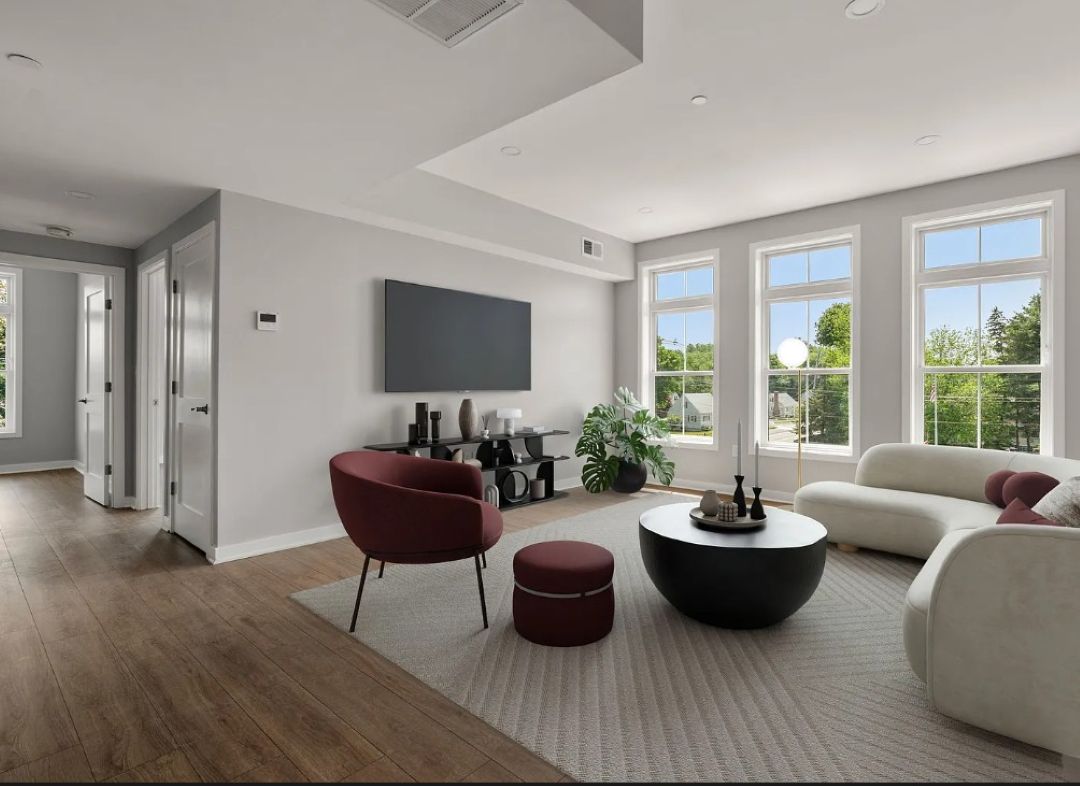 Bright living room with light colored walls, wood-look flooring, and large windows. Features a cream-colored sofa, burgundy accent chair and ottoman, black round coffee table, and wall-mounted television.