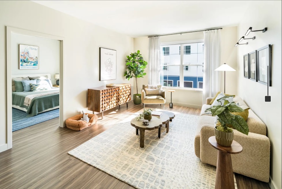 Sunlit apartment living room with a beige sofa, round coffee table, area rug, and large windows.