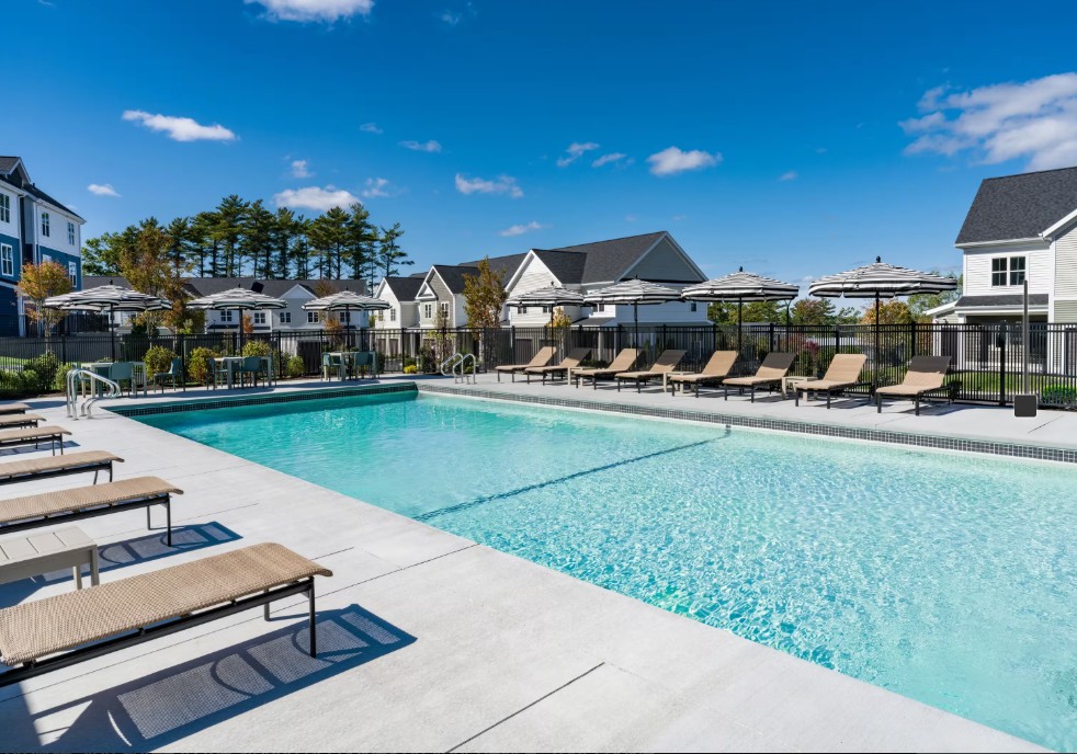 Large outdoor swimming pool surrounded by lounge chairs, umbrellas, and apartment-community buildings.